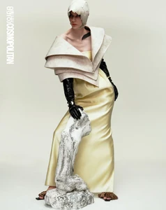 Model in architectural yellow gown and structured sculptural headpiece leaning on a textured white prop against a plain light background.