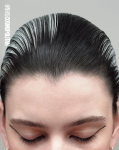Close-up portrait of a model with slicked-back dark hair featuring white streaks against a plain grey background.