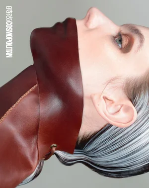 Close-up profile portrait of a model wearing a structured brown leather garment covering the lower face against a grey studio backdrop.
