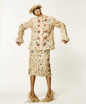 Model wearing a textured straw fringe outfit and matching headpiece against a plain white background