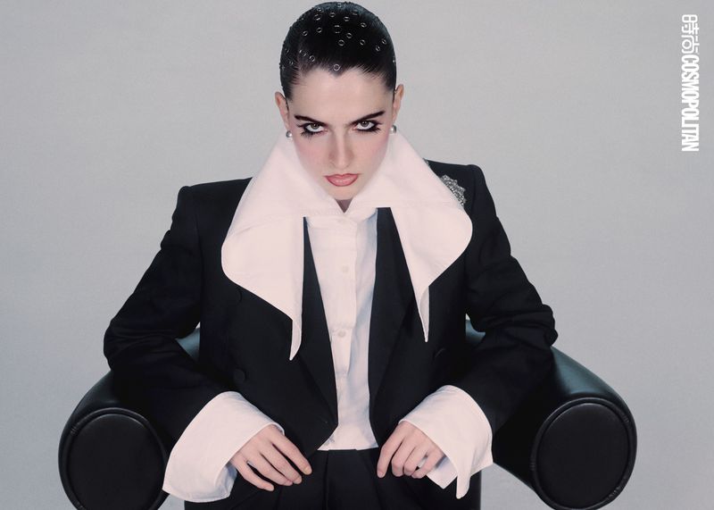 Portrait of a woman with dark hair and dramatic makeup, wearing a black suit and white collared shirt, sitting against a gray background.