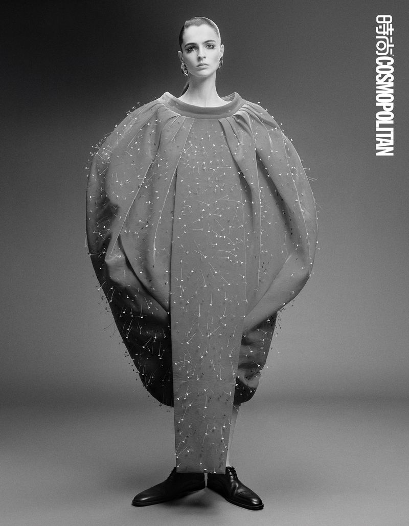 A full-length studio shot showcases a slender female model in an avant-garde, coral gown with spherical accents, set against a muted grey backdrop.