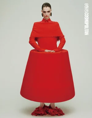 Model wearing a structured red dress with a wide cylindrical skirt and ruffled shoes against a plain white background.