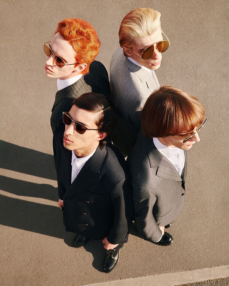 Overhead shot of four people in suits and sunglasses standing back to back in an outdoor setting with neutral ground in the background.