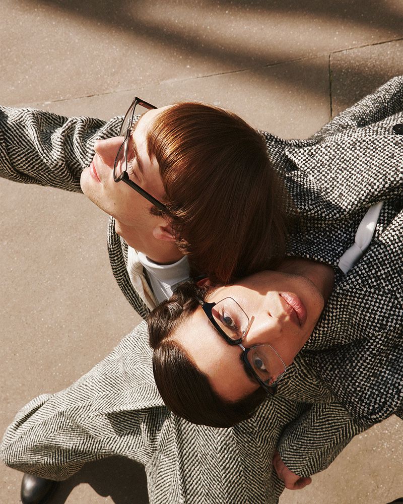 Two people in tweed coats and eyeglasses pose for an eye-level shot on an outdoor urban surface with sunlight and shadows.