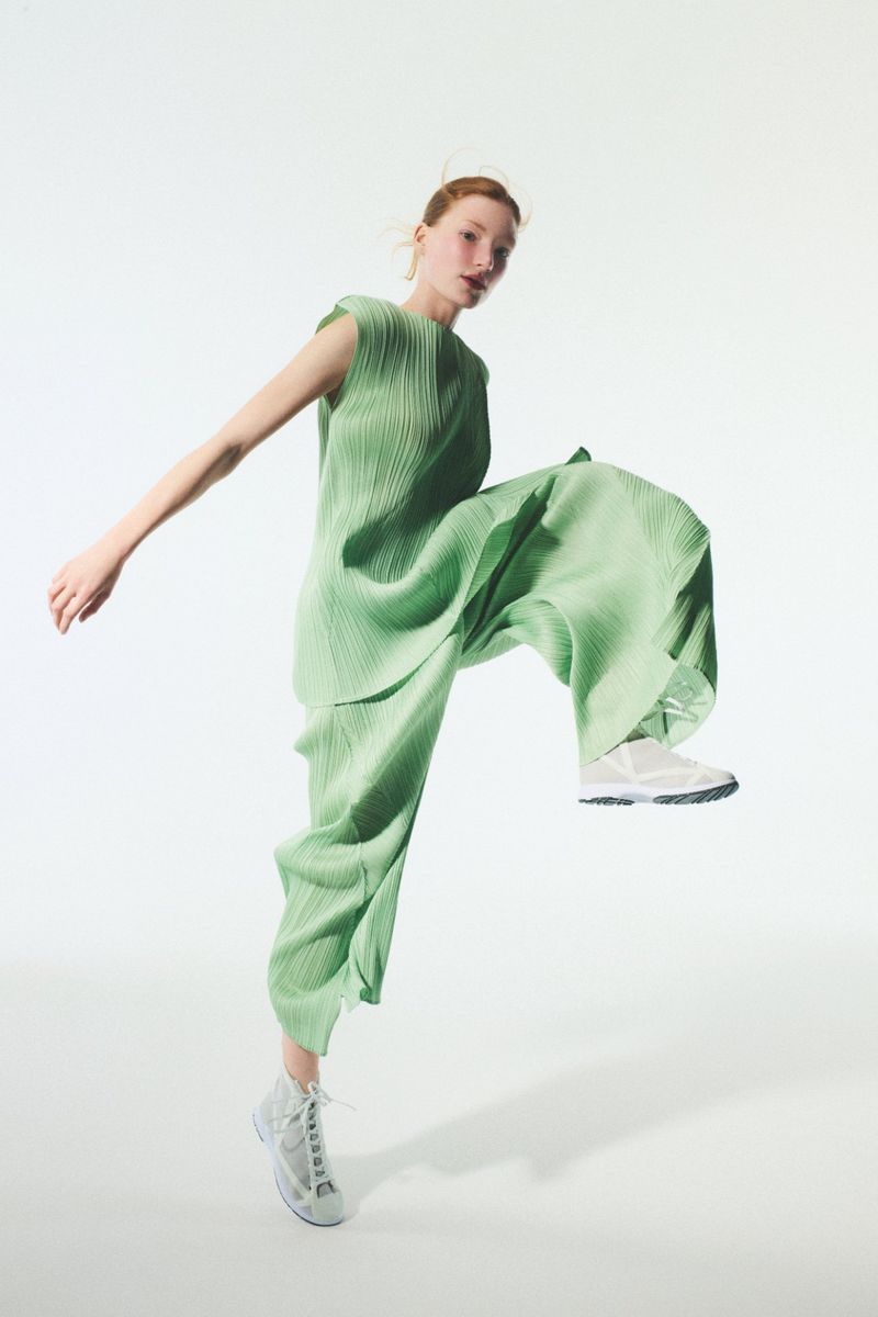 Model in a sage green pleated sleeveless top and matching pants jumping against a white background.
