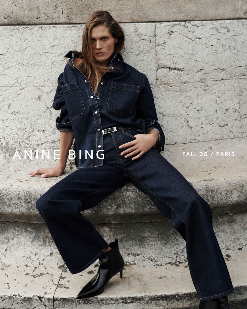Model Malgosia Bela wearing dark denim shirt and jeans sitting against stone exterior for Anine Bing Fall 2026 campaign.