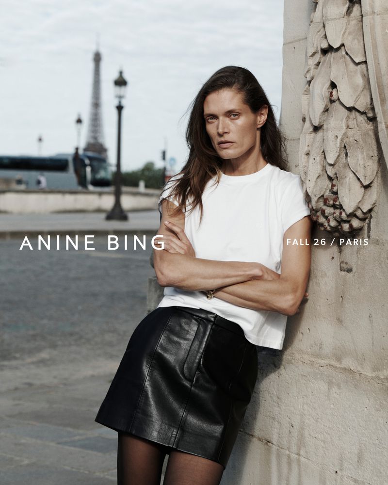 Model Malgosia Bela leaning against an architectural stone pillar in Paris with the Eiffel Tower in the background wearing a white t-shirt and black miniskirt.