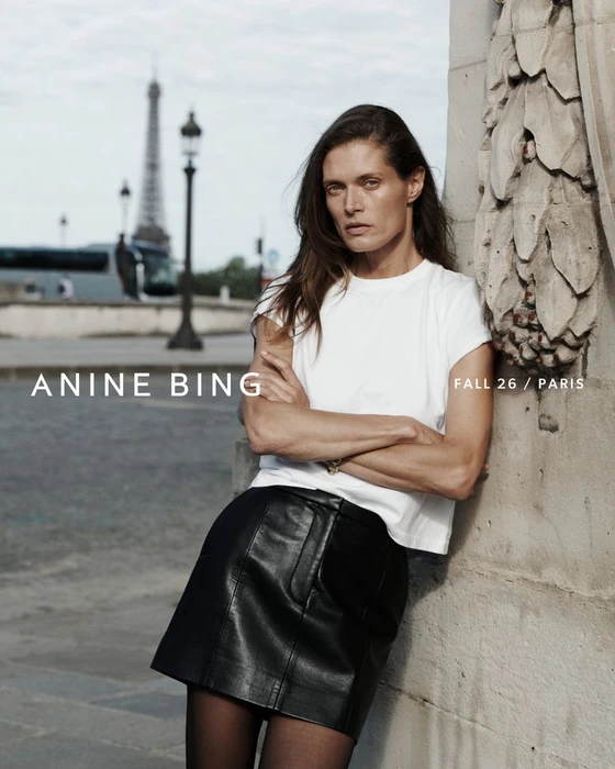Model Malgosia Bela leaning against an architectural stone pillar in Paris with the Eiffel Tower in the background wearing a white t-shirt and black miniskirt.