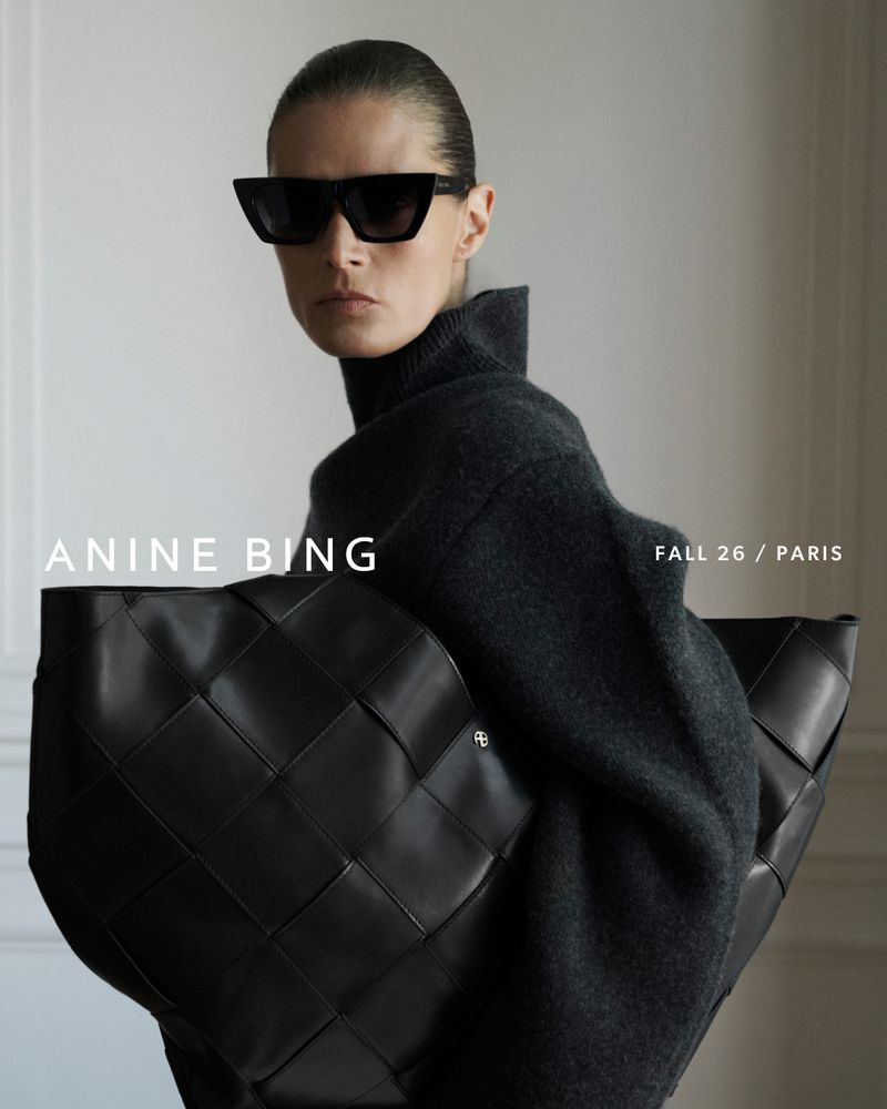 Model wearing dark sunglasses and a charcoal turtleneck holding a black woven leather tote bag for Anine Bing Fall 2026.