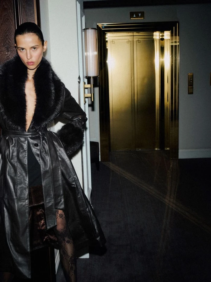 Model in a black leather coat with faux fur collar standing in a hotel hallway near gold elevator doors.
