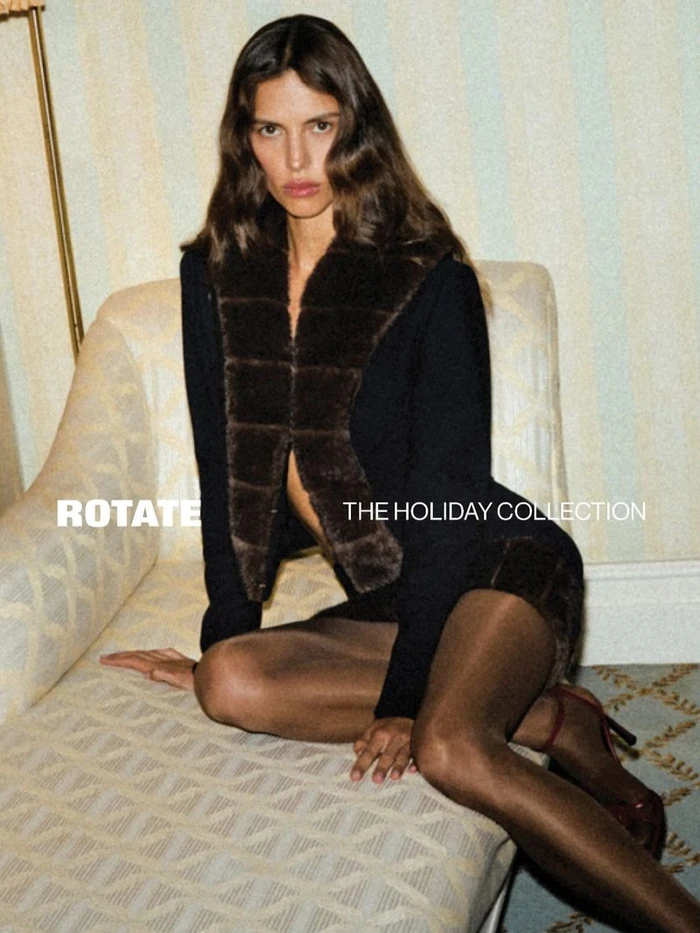 Model seated on a patterned sofa wearing a dark jacket with a fur collar for Rotate