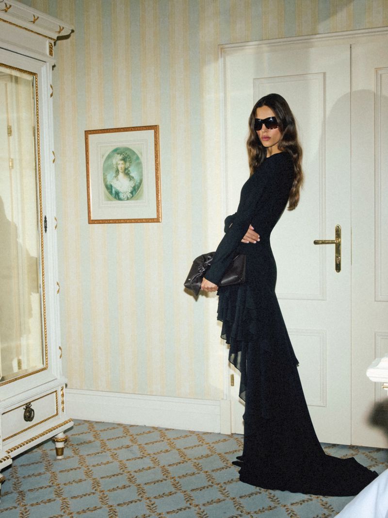 Model in a long black dress and sunglasses holding a clutch bag in a room with striped wallpaper and patterned carpet.