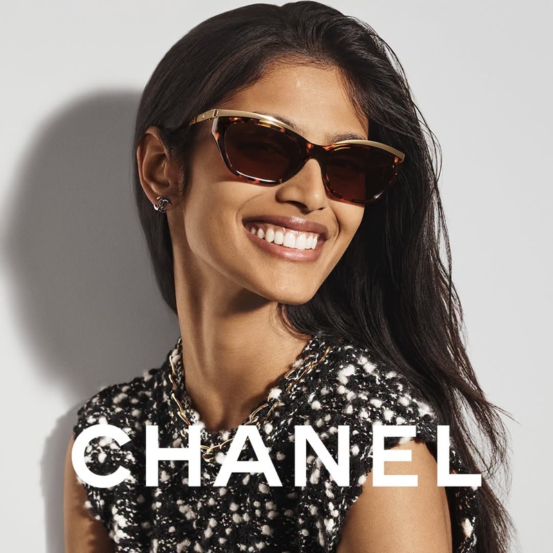 Close-up portrait of a smiling woman wearing tortoiseshell sunglasses and a textured tweed top against a plain white background.