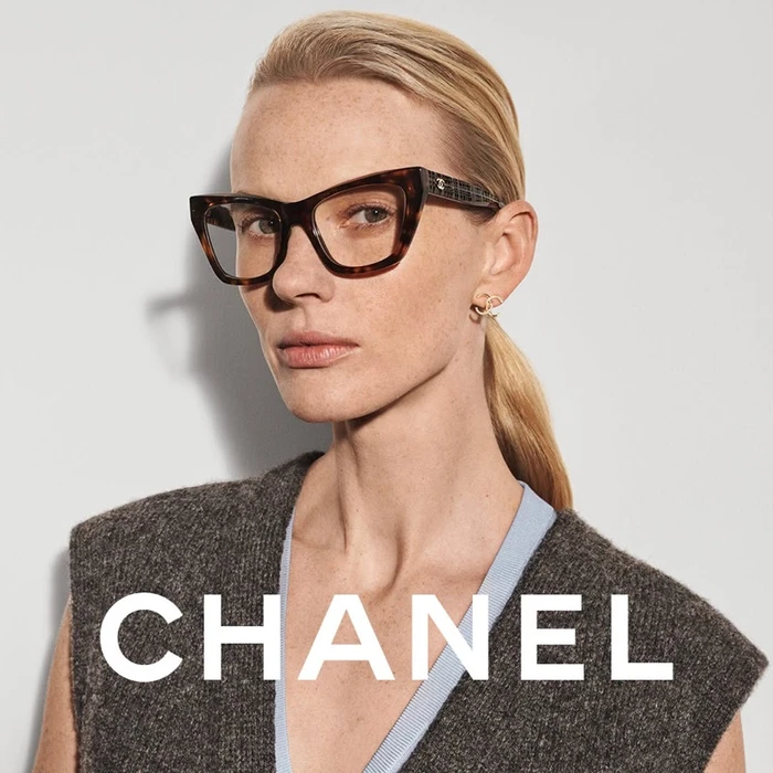 Close-up portrait of a blonde model wearing tortoiseshell eyeglasses and a grey vest against a white background for Chanel.