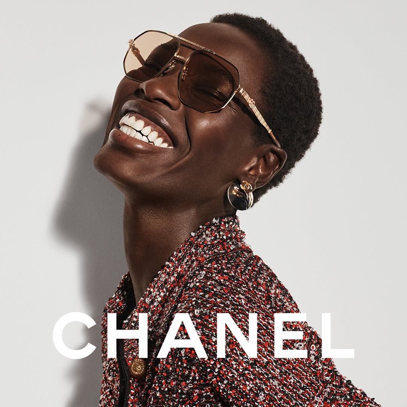 Close-up portrait of a Black woman smiling while wearing tinted aviator sunglasses and a red tweed jacket against a plain background.