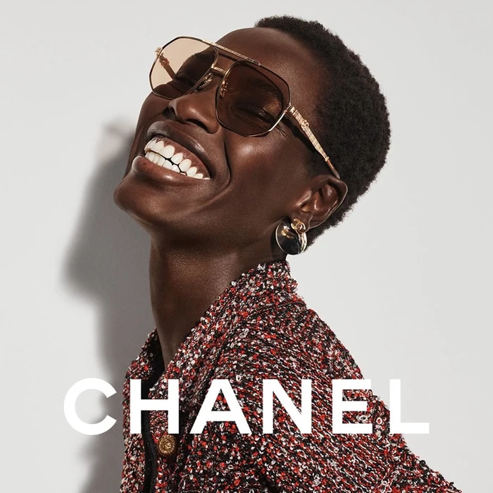 Close-up portrait of a Black woman smiling while wearing tinted aviator sunglasses and a red tweed jacket against a plain background.