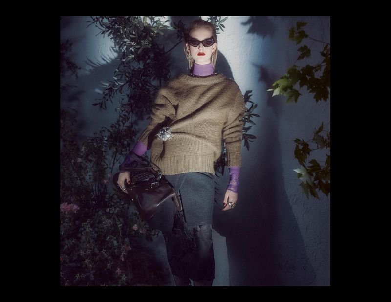 Model in dark sunglasses wearing a textured brown sweater and holding a dark handbag against foliage