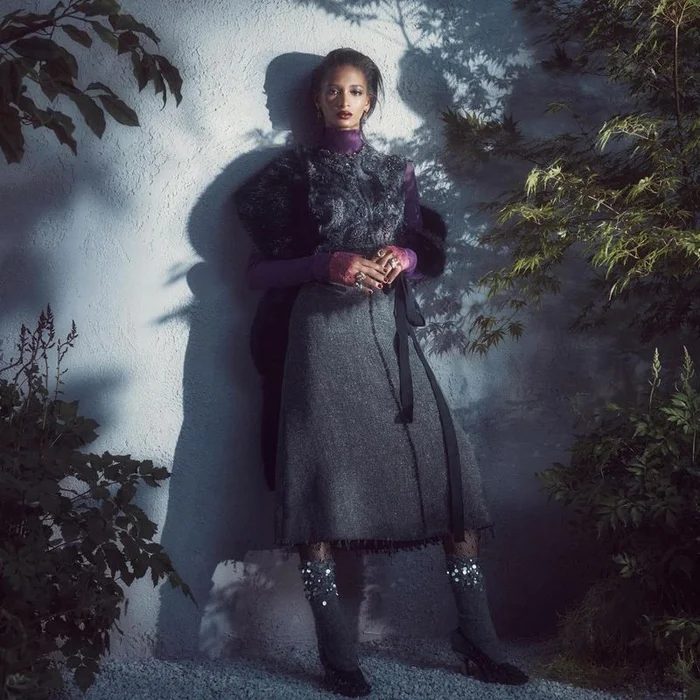 Model leaning against a textured wall wearing a grey skirt, patterned top, and dark faux fur wrap with foliage casting shadows.
