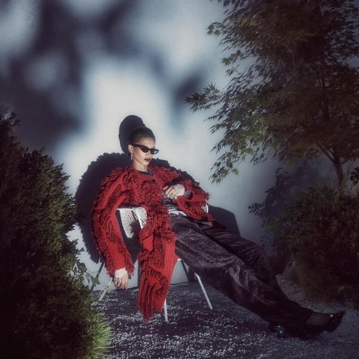 Model wearing dark sunglasses and a red textured coat seated on a white chair against a wall with tree shadows