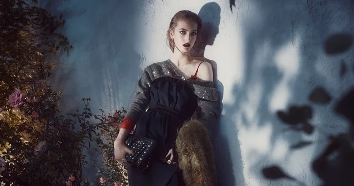 Model leaning against a textured wall with floral bushes, holding a dark studded handbag and wearing a black dress and sequined sweater.