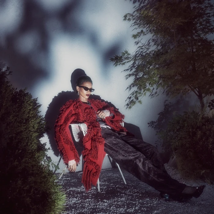 Rihanna wearing sunglasses and a textured red coat reclining in a chair outdoors at night under dramatic shadows.
