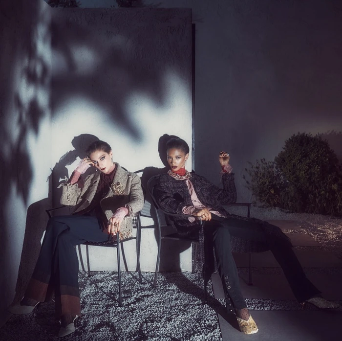 Two women seated in outdoor chairs against a white wall with harsh leaf shadows for Zara Twilight Roses