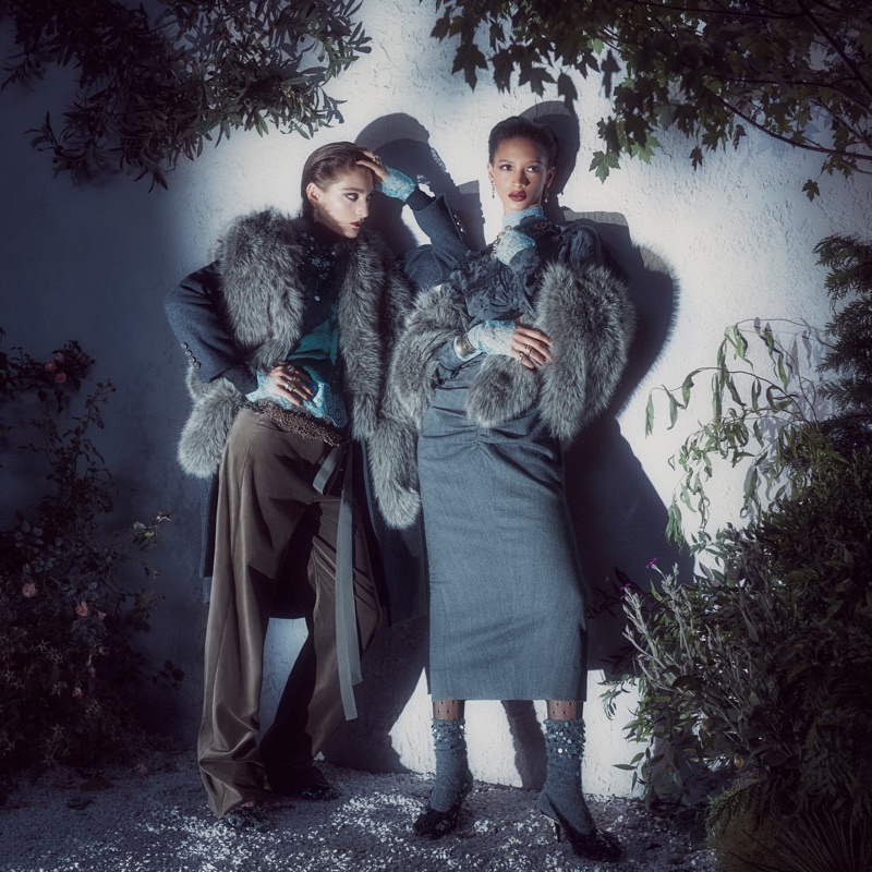 Two models in fur-trimmed coats and layered autumn apparel posing outdoors against a pale wall at night.