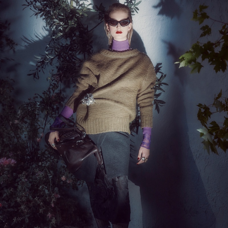 Model wearing dark sunglasses, knit sweater, and holding a handbag outdoors at night under dramatic lighting.