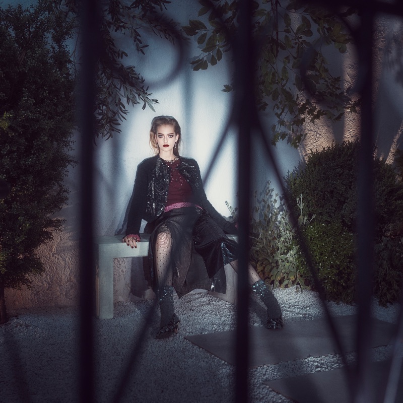 Model seated on a stone bench in a dim garden behind dark wrought-iron bars wearing a dark coat and sheer skirt.
