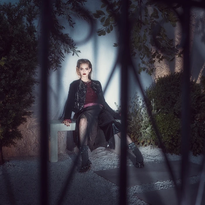 Model seated on a stone bench in a dim garden behind dark wrought-iron bars wearing a dark coat and sheer skirt.