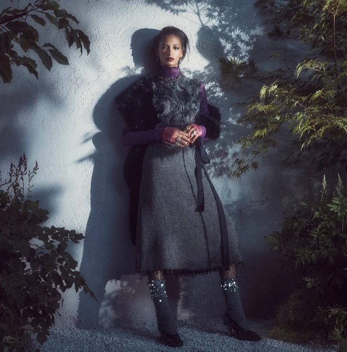 Model in grey skirt and dark patterned top leaning against a textured wall surrounded by foliage