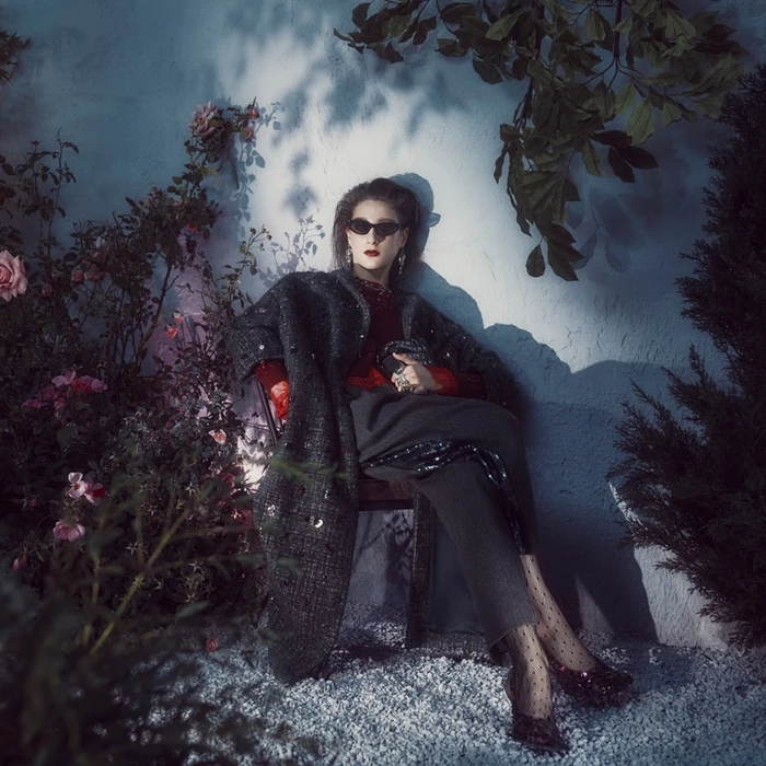 Model seated outdoors wearing sunglasses and a textured dark coat amidst climbing pink roses and shadows under moonlight.