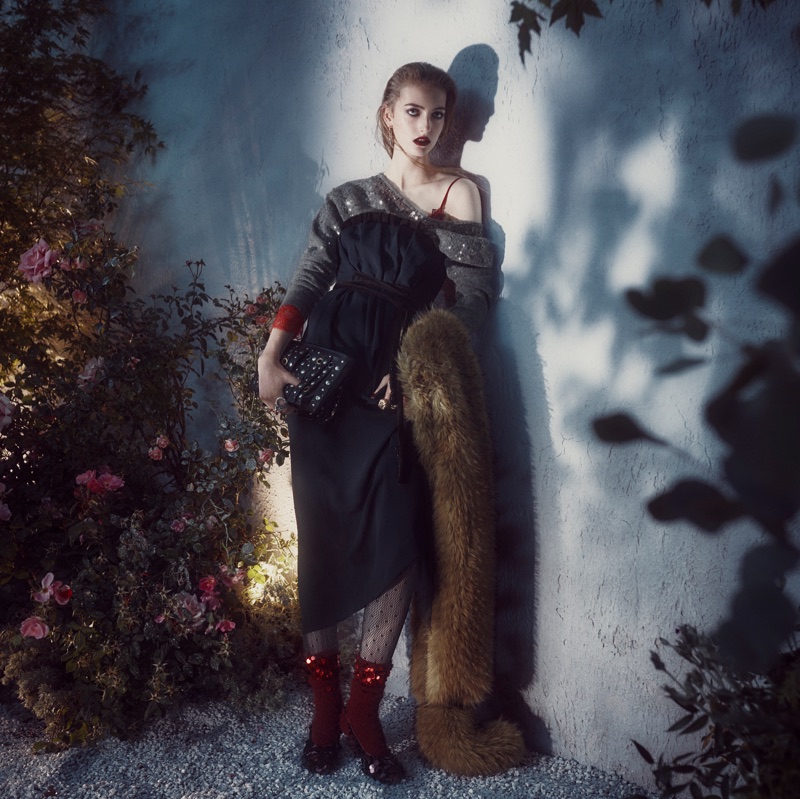 Model leaning against a textured wall next to rose bushes wearing a dark dress, textured sweater, and holding a handbag.