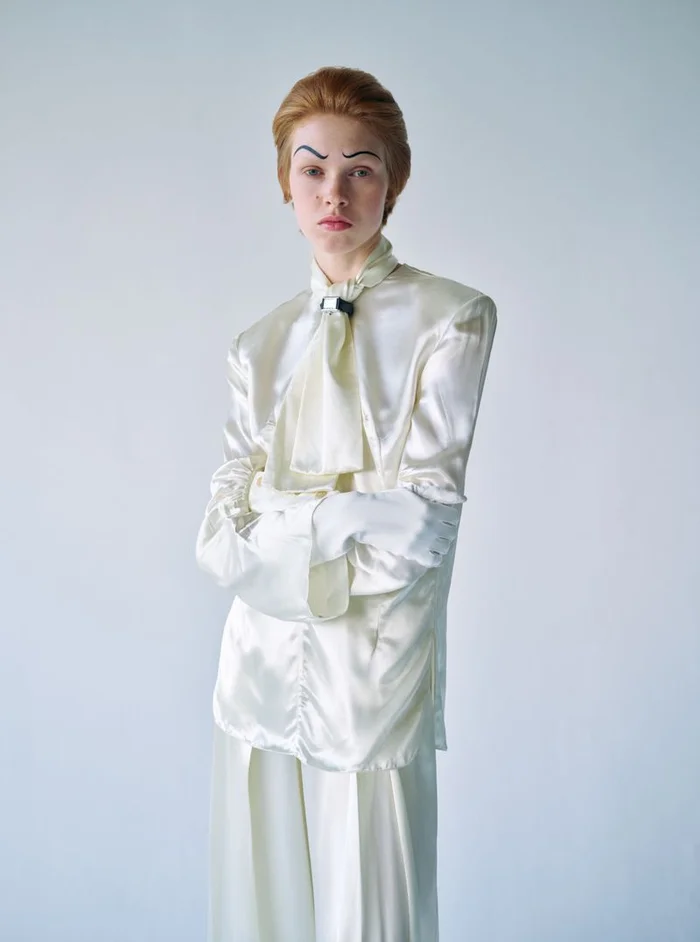 Model with styled arched eyebrows wearing a shiny cream silk blouse and matching trousers against a plain white background