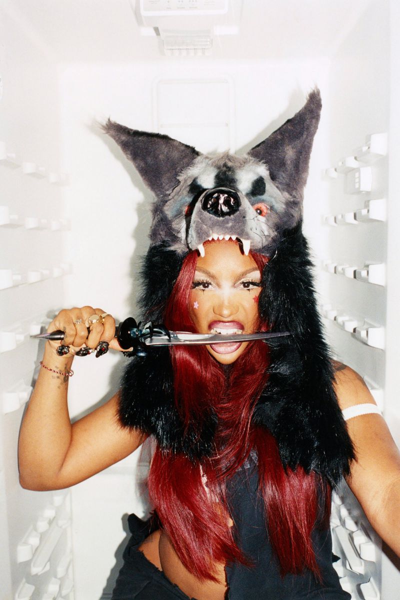Woman with red hair wears a furry wolf hat while biting down on a knife in a white refrigerator, creating an edgy and fierce portrait.