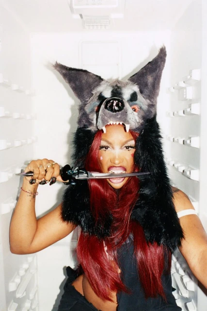 Woman with red hair wears a furry wolf hat while biting down on a knife in a white refrigerator, creating an edgy and fierce portrait.