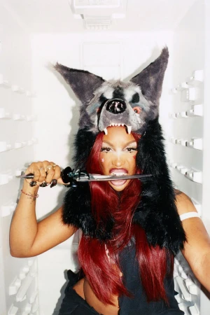 Woman with red hair wears a furry wolf hat while biting down on a knife in a white refrigerator, creating an edgy and fierce portrait.