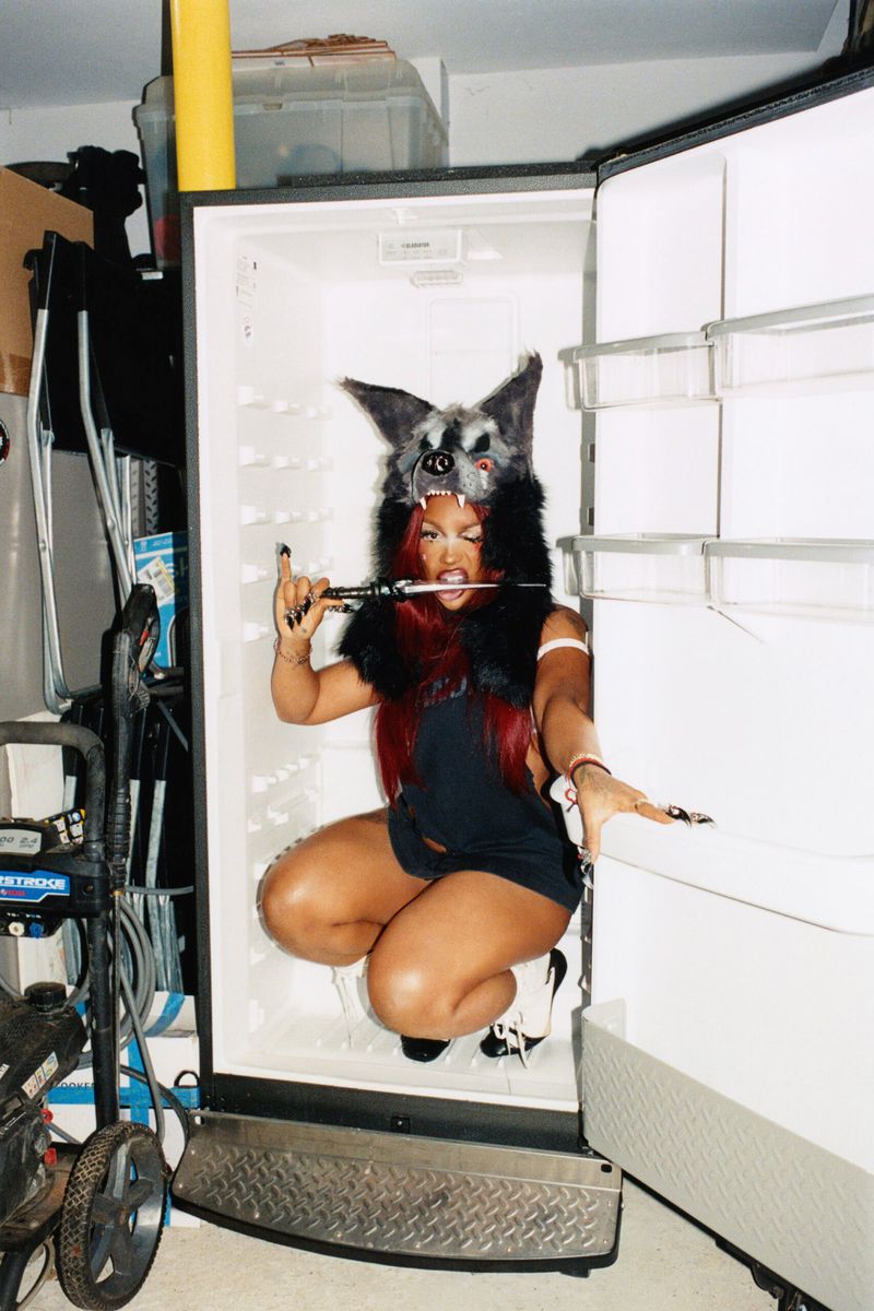 Adult woman in a wolf hat crouches in an empty refrigerator holding a knife in her mouth, creating an eerie image in a garage setting.