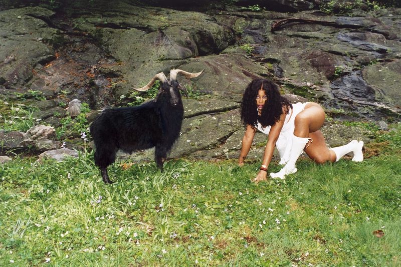 Adult female poses with a black goat in a meadow with a rocky background, wearing a white dress and white boots.