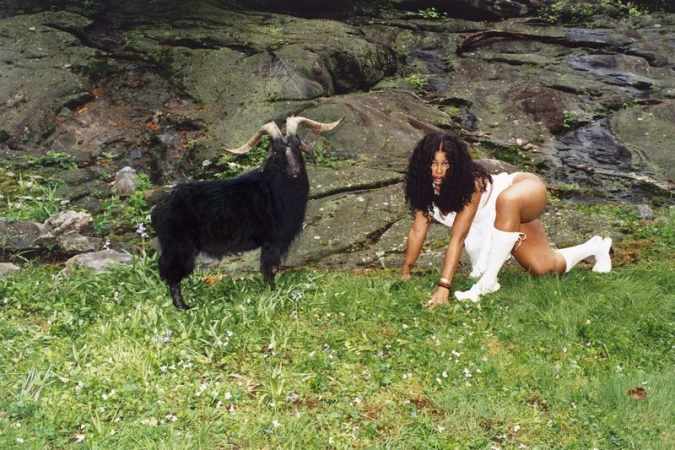 Adult female poses with a black goat in a meadow with a rocky background, wearing a white dress and white boots.