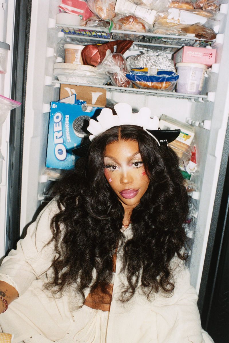 Adult female with dark curly hair in white top and headpiece sitting in front of a fridge filled with food containers and Oreo box.
