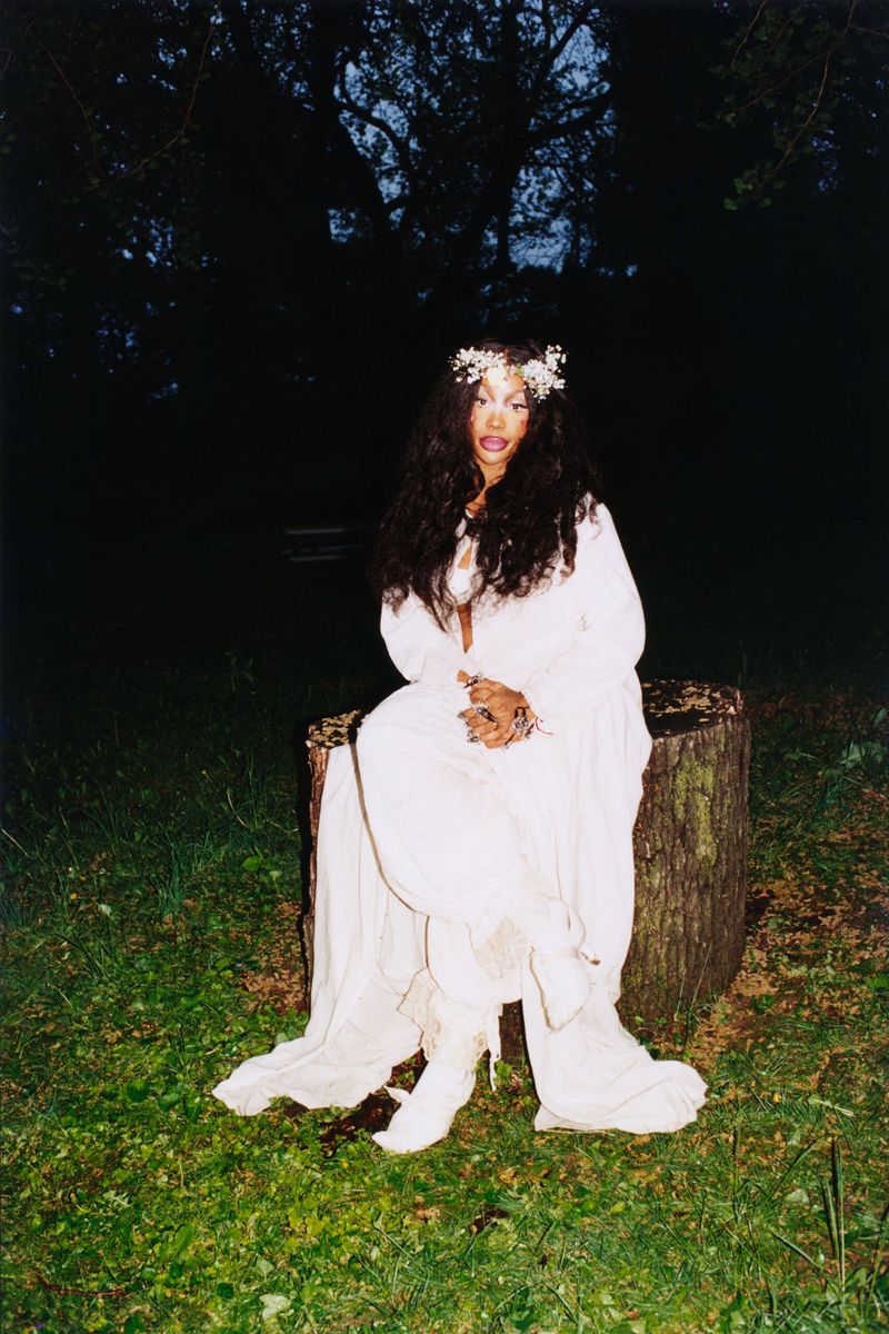 A woman in a white dress sits on a tree stump outdoors in a lush green field under a dark tree canopy and dim lighting.