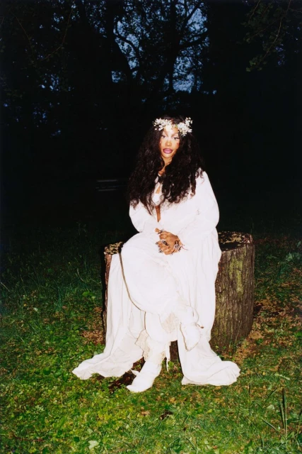 A woman in a white dress sits on a tree stump outdoors in a lush green field under a dark tree canopy and dim lighting.