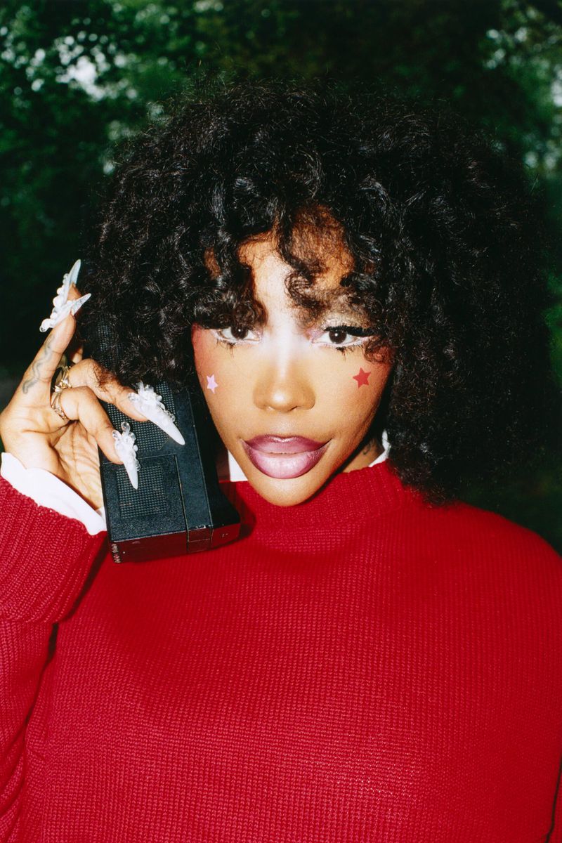 Close-up of a young African-American woman with curly hair holding a black rectangular object, wearing red sweater, with red star makeup.