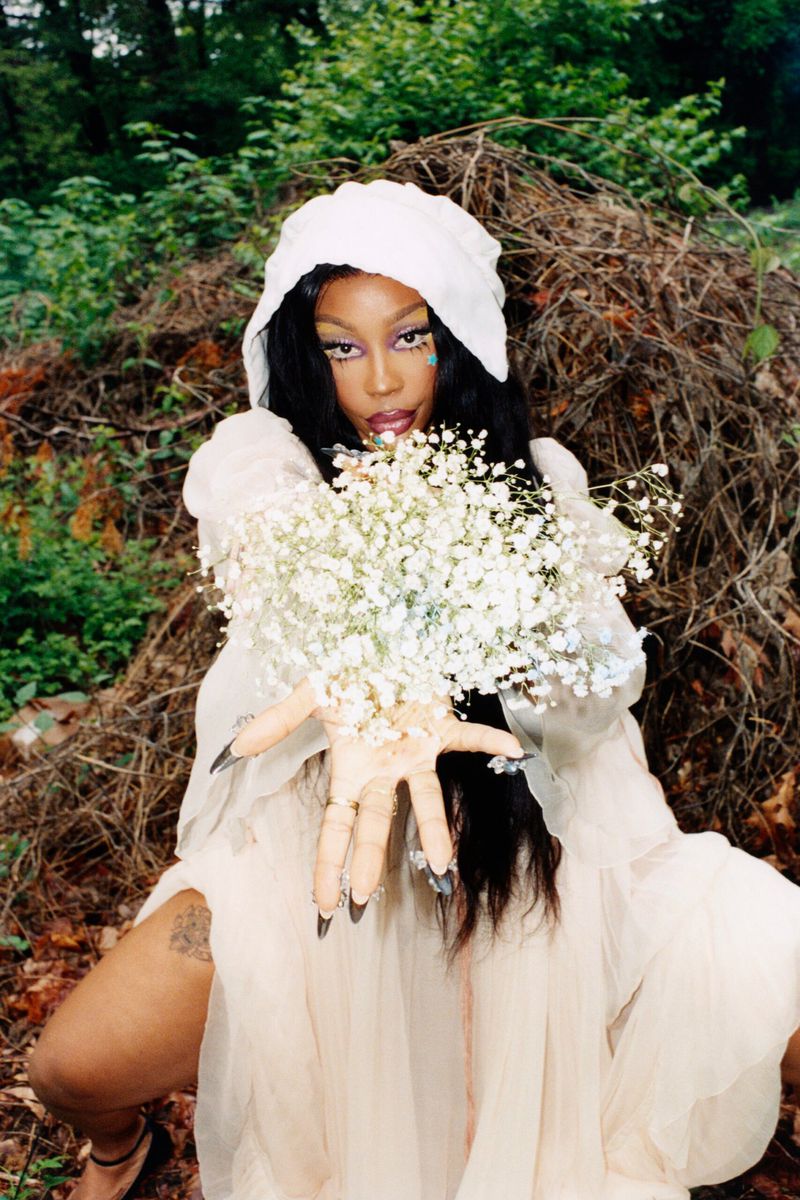 A fair-skinned woman in a draped cream-colored hooded gown poses with white baby's breath flowers in a wooded outdoor setting.