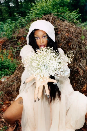A fair-skinned woman in a draped cream-colored hooded gown poses with white baby's breath flowers in a wooded outdoor setting.