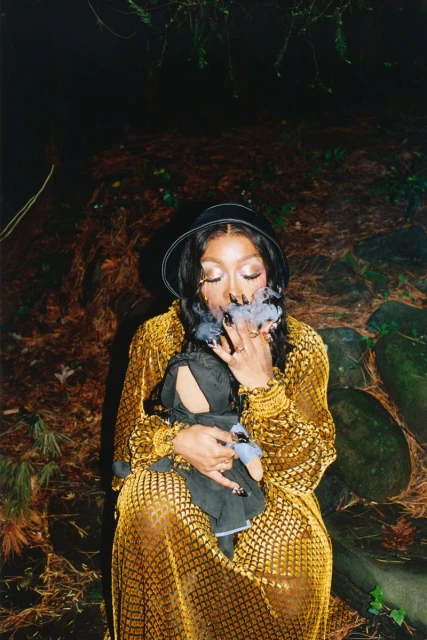 Woman in gold dress holds doll while smoke surrounds her face in outdoor portrait. She wears a hat, stands in a wooded area.