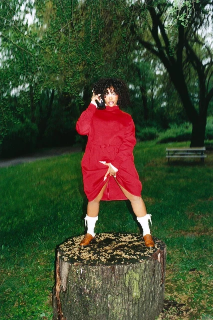 Woman in red dress and white boots stands on a tree stump in a grassy park with trees, talking on a vintage-style phone.