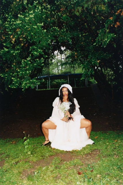Woman in a white dress and bonnet sits in the grass with wildflowers, in a wooded area under leafy trees.